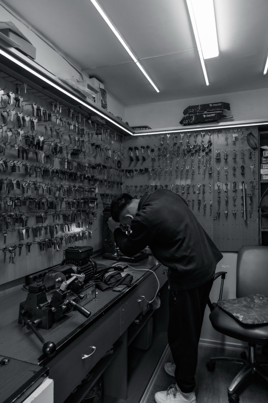 Locksmith focused on a key-cutting machine at the bench, wall of key blanks in the background